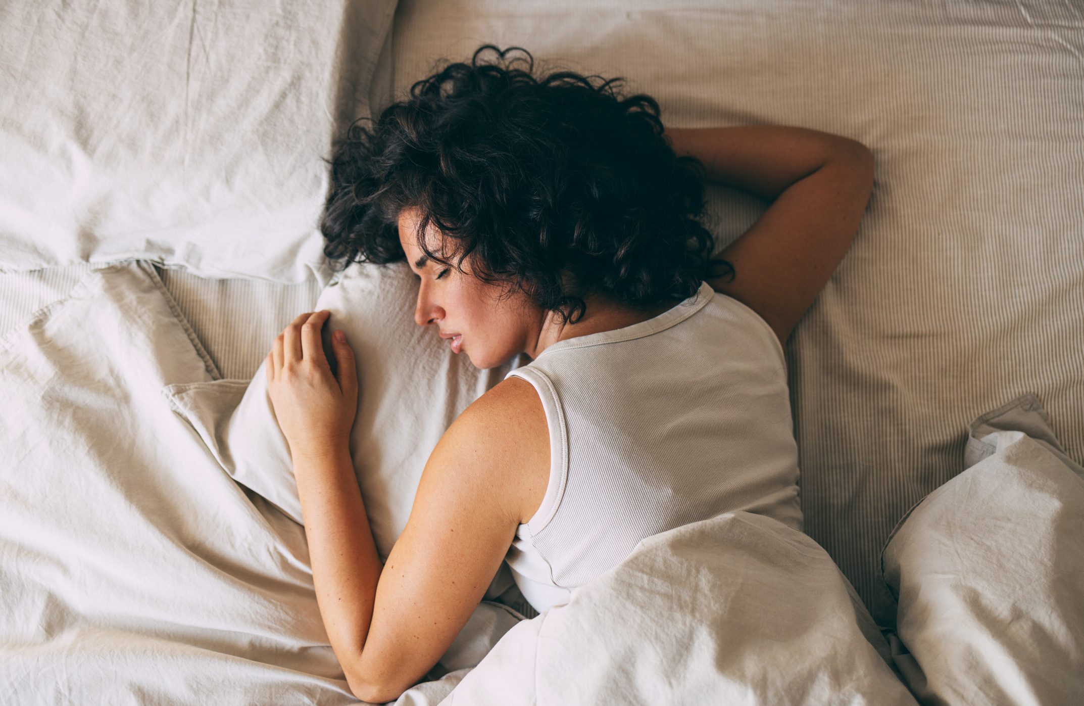 A woman with curly hair resting peacefully on a bed in a white tank top.