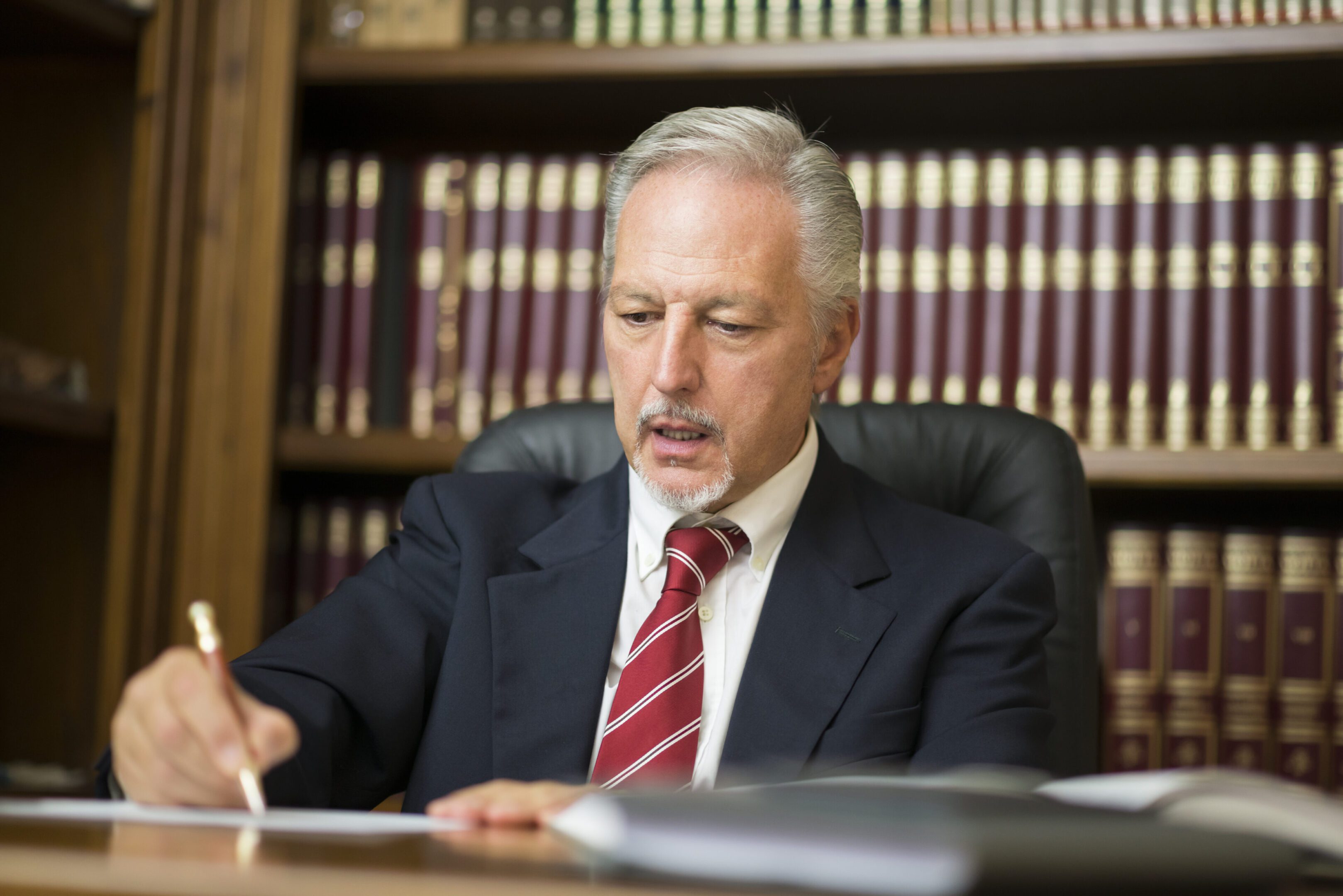 A man in a suit and red striped tie reading at a desk with bookshelves behind him.