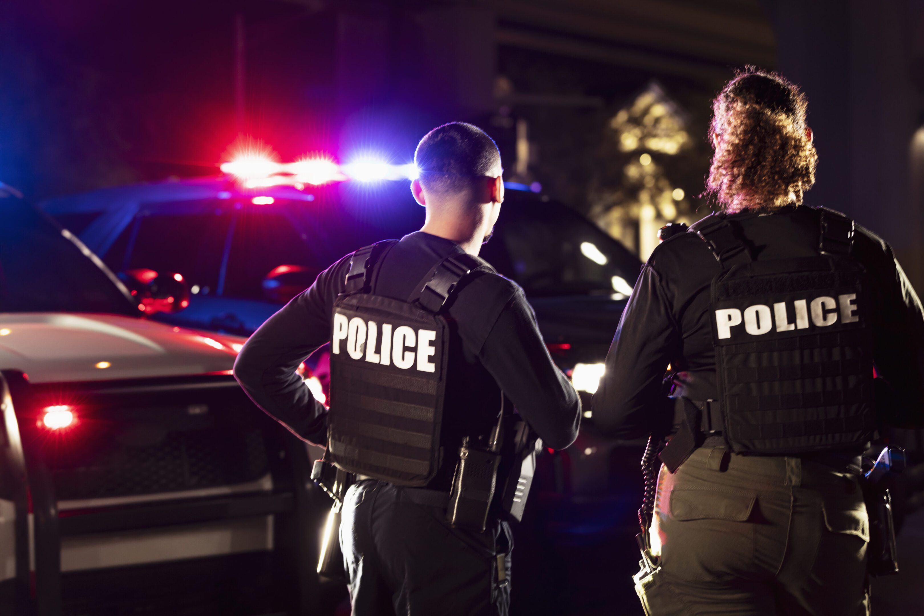 Two police officers standing near a patrol car with flashing lights at night.