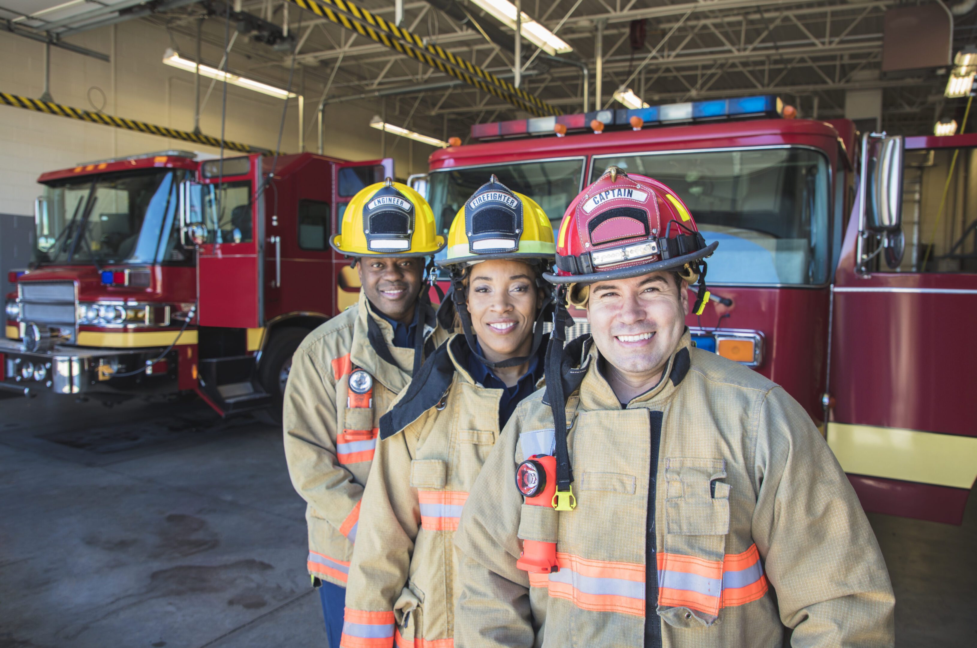 Three firefighters in uniform smiling inside a fire station.